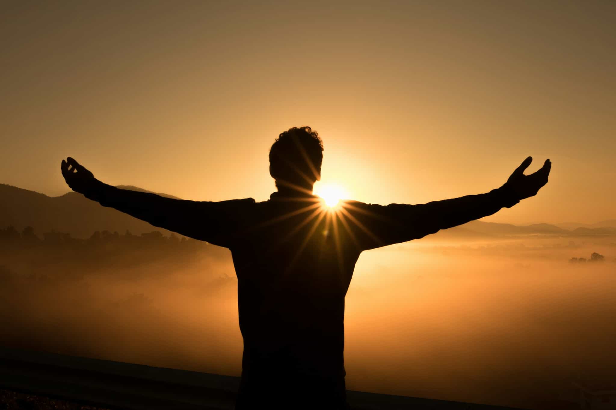 Silhouette mit ausgestreckten Armen vor goldenem Sonnenaufgang